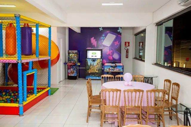 Salão com mesa redonda rosa, cadeiras de madeira, escorregador laranja e fliperamas