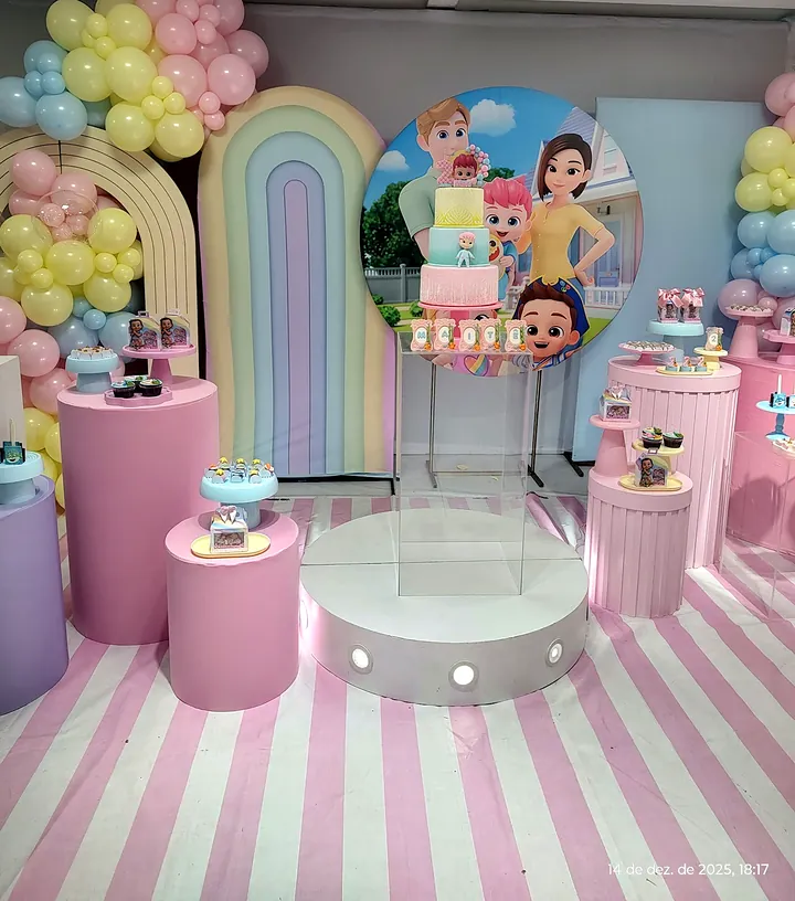 Salão decorado em tons de rosa, com arcos, balões pastel, bolo sobre a mesa branca e tapete listrado de rosa e branco